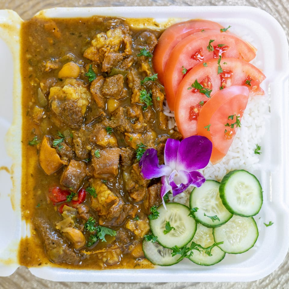 Hazel's Jamaican curry goat with rice, sliced tomatoes, cucumbers, and a purple orchid in a takeout container.